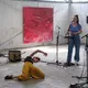Vier Personen mit heller Haut in einer Industriehalle bei einer Performance. Links spielt eine Frau Gitarre, in der Mitte singt eine Frau am Mikrofon, rechts steht eine weitere Frau an einem Mikrofon. Davor tanzt eine Frau auf dem Boden. Im Hintergrund hängen Kunstwerke.