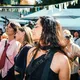 Eine scheinbar junge Frau mit heller bis olivfarbener Haut und langen dunklen Haaren steht mit geschlossenen Augen in einer Menschenmenge auf einem Festival. Im Hintergrund sind weitere Personen unterschiedlicher Hauttöne, Sonnenschirme und Wimpel zu sehen.