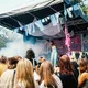Eine scheinbar weibliche Person mit dunkler Haut singt auf einer Festivalbühne ins Mikrofon. Vor der Bühne steht ein Publikum mit Personen verschiedener Hauttöne. Nebel, Bühnendekoration und bunte Stoffbänder schaffen eine lebendige Konzertatmosphäre.